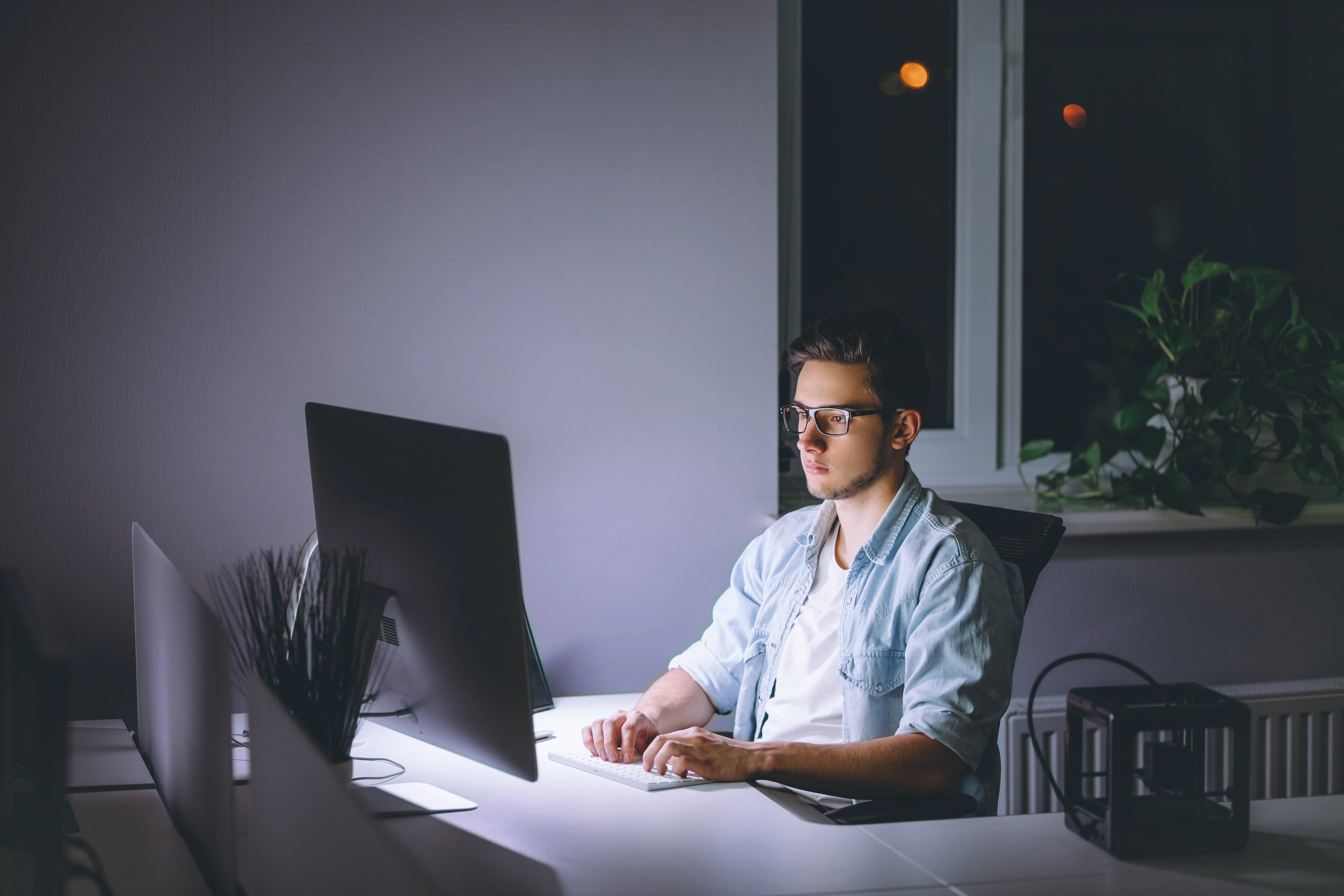 Young man working on computer at night in dark office. The designer works in the later time. A young man sits at the computer